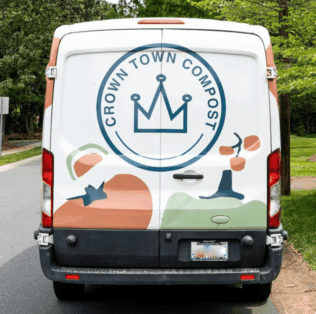 The back of a white Crown Town Compost van featuring a large blue crown logo and abstract shapes of fruits and vegetables, parked on a tree-lined street.