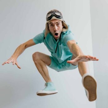 A man in a teal outfit and aviator cap with goggles jumps mid-air with an animated expression, reaching his arms forward.