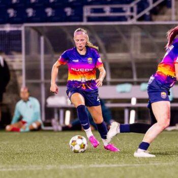 A female soccer player in a colorful jersey dribbles the ball on the field during a match, with another player beside her.