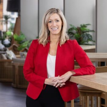 A woman in a red blazer and white blouse stands smiling in a modern indoor space with light wood furniture and green plants in the background.