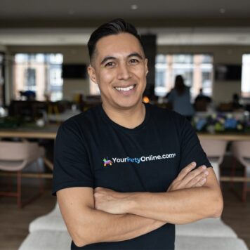 A smiling man stands with arms crossed wearing a black t-shirt that says “YourPartyOnline.com” in a casual indoor setting.
