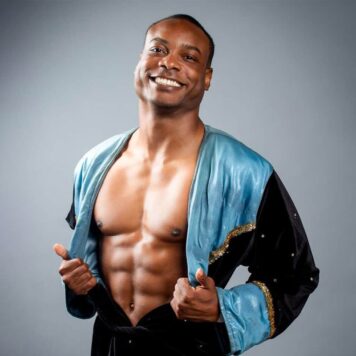 A smiling man poses in an open wrestling robe with shimmering blue and black fabric, against a gray background.