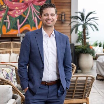 A man in a blue suit smiles while standing on a stylish outdoor patio, with tropical plants and vibrant artwork in the background.