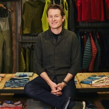 A man in a dark button-up shirt and jeans sits on a display table surrounded by folded clothes and colorful outdoor jackets in a retail store.