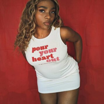 A woman poses confidently in front of a red backdrop, wearing a white sleeveless dress with the words 