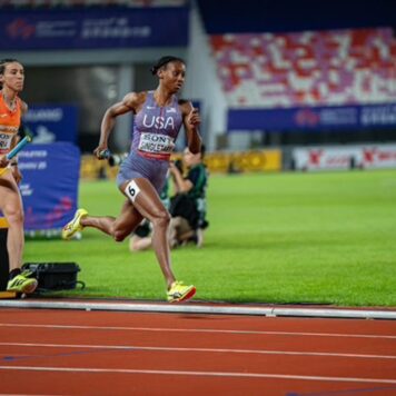 A track athlete runs a relay race holding a baton, competing alongside another runner on a professional track.