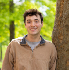 A UNC Charlotte student leans against a tree in a wooded area of campus filled with bright green foliage.