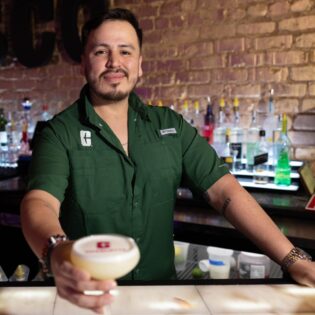 Man in green Charlotte polo shirt looking at the camera and holding out a cocktail.