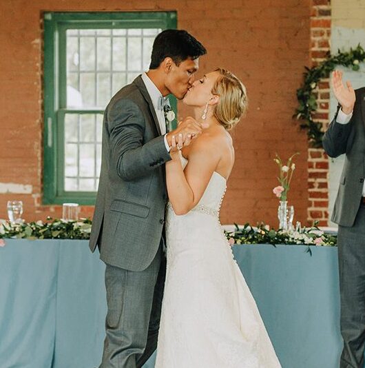 KEVIN-KATE-Niner-Love-Story A bride and groom share a kiss during their wedding ceremony in a brick venue while bridesmaids and groomsmen applaud in the background.