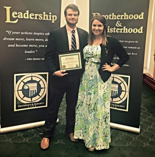 Lauren-Smith-Niner-Love-Story-1 A man holding a certificate stands beside a woman at a fraternity and sorority life awards event, posing in front of banners that read “Leadership” and “Brotherhood & Sisterhood.”