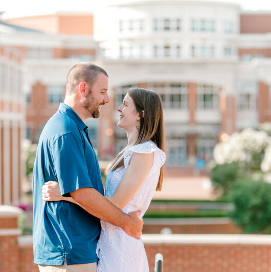 Sarah-Kraft-Niner-Love-Story A couple stands embracing and smiling at each other in front of a brick campus building on a sunny day.