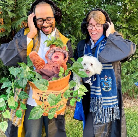 Shane-Taylor-Niner-Love-Story Two adults wearing robes and earmuffs smile while holding a baby dressed as a mandrake in a flowerpot, with a small white dog beside them outdoors.