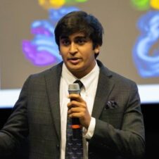 Rishi Misra UNC Charlotte student in a suit speaks on stage with a microphone during a presentation with a colorful screen in the background.