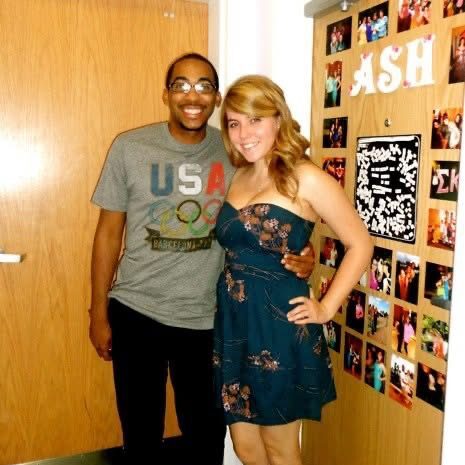 Shane-Taylor-Niner-Love-Story-1 Two college students stand smiling in a dorm room doorway decorated with photos and a name sign, posing together for a casual picture.