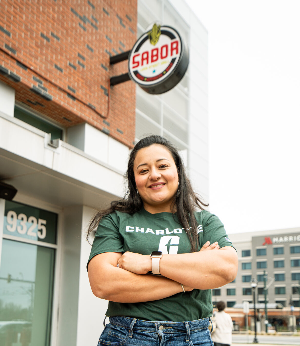 Charlotte_Alumni_Miriam-Espilliat_SABOR Smiling woman in a Charlotte shirt outside Sabor restaurant