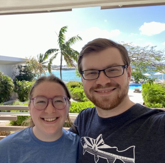 Hayley-Owen-Niner-Love-Story-1 A smiling couple takes a selfie on a balcony overlooking palm trees, a pool, and bright blue ocean water on a sunny day.