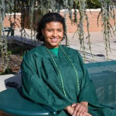 Madison Black Smiling UNC Charlotte graduate sits on a campus bench in green regalia, under drooping tree branches.