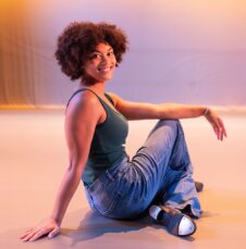 Commencement_Niya-Kerr Charlotte student in tank top and jeans poses seated on the floor under warm, colorful lighting.