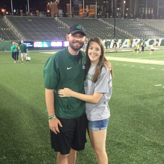 Sarah-Kraft-Niner-Love-Story-1 A couple stands arm in arm on a football field at night, with stadium seating and a campus building visible in the background.