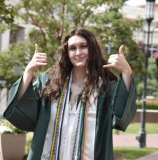 Commencement_Anna_Shimota Graduate in green gown gives a double thumbs-up on campus, wearing honor cords.