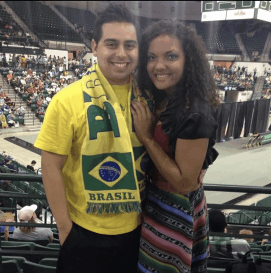 Sierra-Flores-Niner-Love-Story A couple poses together inside a crowded arena, with the man wearing a yellow Brazil shirt and green scarf.