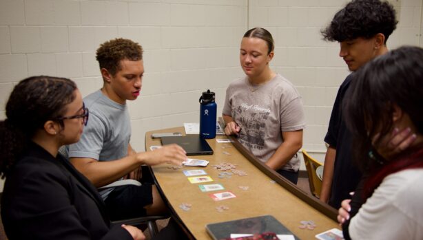 Students Playing PLACES Game in Classroom A group of college students gathered around a curved classroom desk, playing the board game PLACES. Game cards and tokens are spread out on the table, and a laptop and water bottle are also visible. The classroom has white brick walls and a clock on the wall.