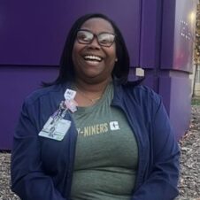Nataysha Addison UNC Charlotte alumna wearing scrubs and a Niners shirt sits smiling outside near a purple building.