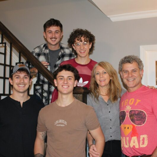 Sharon-Williams-Niner-Love-Story-2 Six family members pose together indoors near a staircase, smiling for a group photo in a warmly lit home.