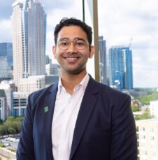 Commencement_Yash-Tadimalla Professionally dressed man smiles in front of large windows overlooking a modern city skyline.