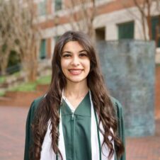 Joy-Kamel UNC Charlotte graduate wearing green regalia and cords, standing outside on campus with a smile.