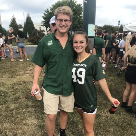 Katerina-Lantz-Niner-Love-Story-1 Two fans wearing Charlotte green apparel pose together at a crowded outdoor tailgate, smiling and holding drinks.