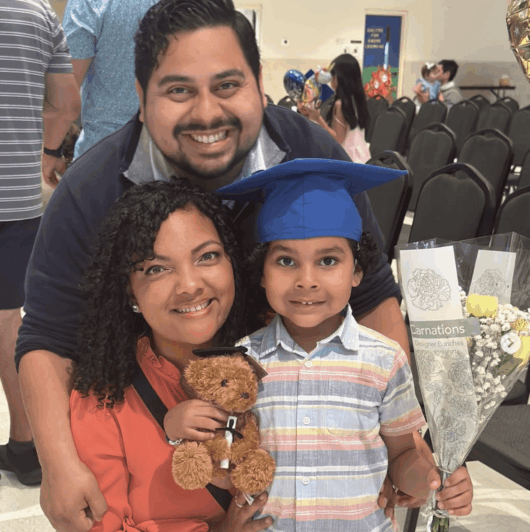 Sierra-Flores-Niner-Love-Story-1 A young child wearing a blue graduation cap stands between two smiling adults, holding a teddy bear and a bouquet of carnations at a celebration event.