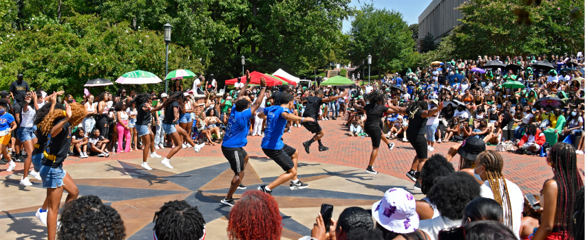 A fraternity and sorority stepping at the NPHC yard show in the Star Quad. They are surrounded by a crown of people cheering them on.