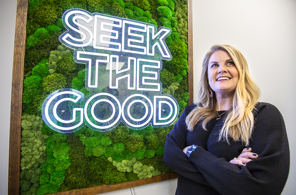 Torrie Savage in front of a piece of moss art displaying the company's mantra: Seek The Good
