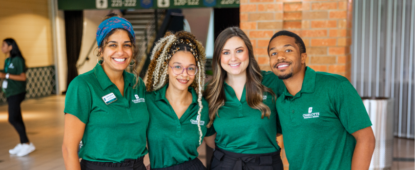 Four staff members in green polos standing and smiling for the camera in the Barnhardt Student Activity Center.