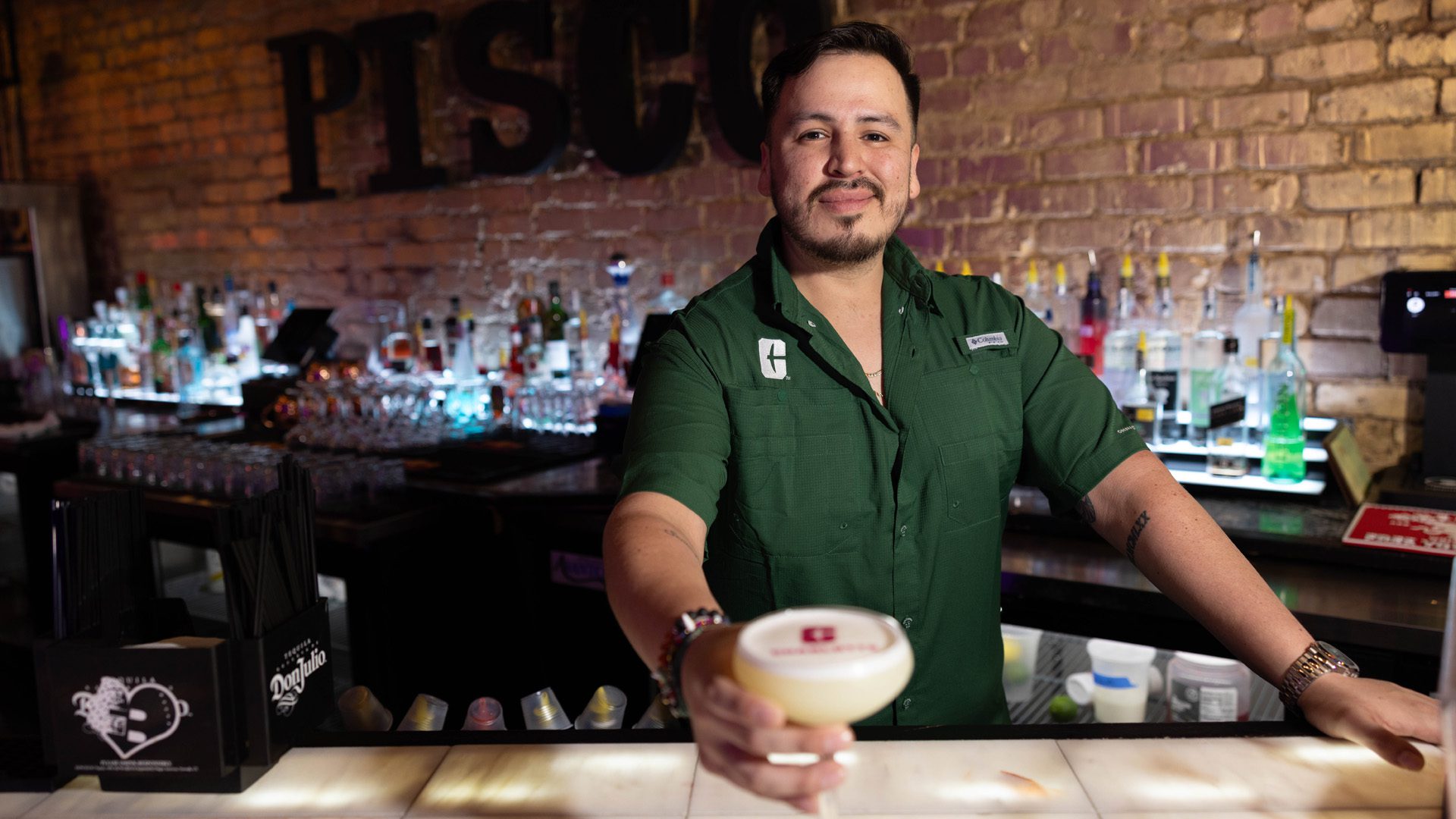 Pisco 1920x1080 Man in green Charlotte polo shirt looking at the camera and holding out a cocktail.