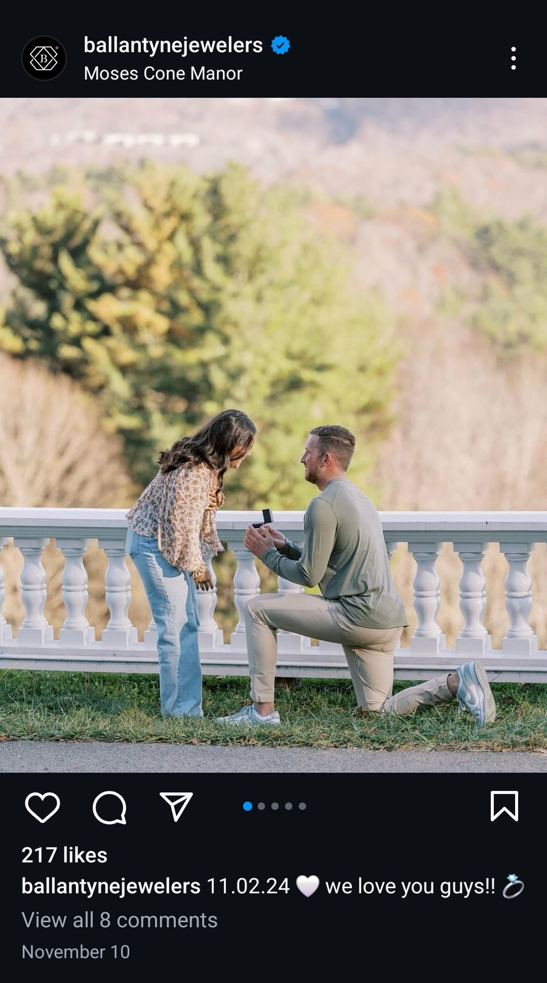 A man kneels and proposes to a woman outdoors by a white balustrade.