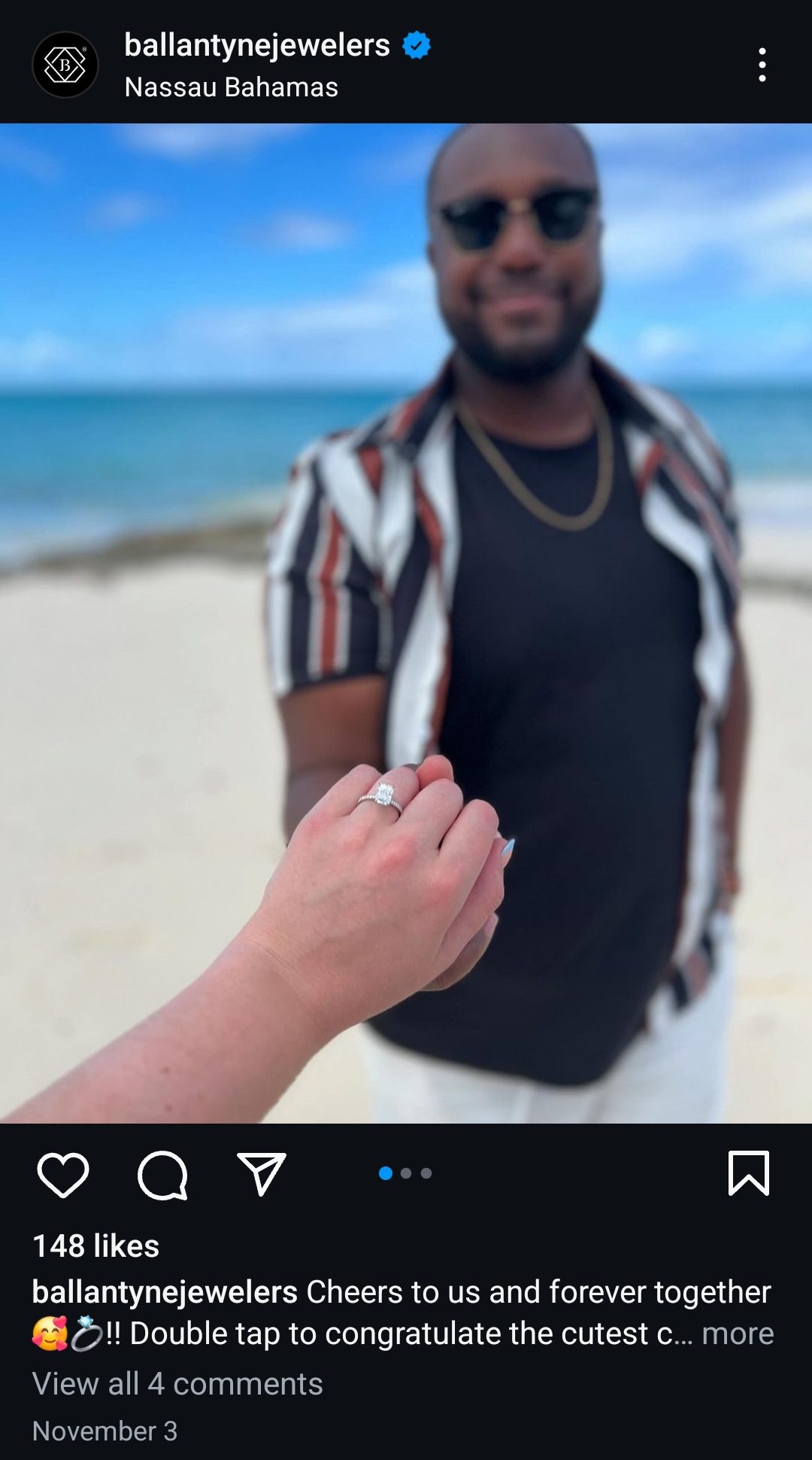 A hand with an engagement ring is held up in the foreground, with a person smiling in the blurred background on a beach.