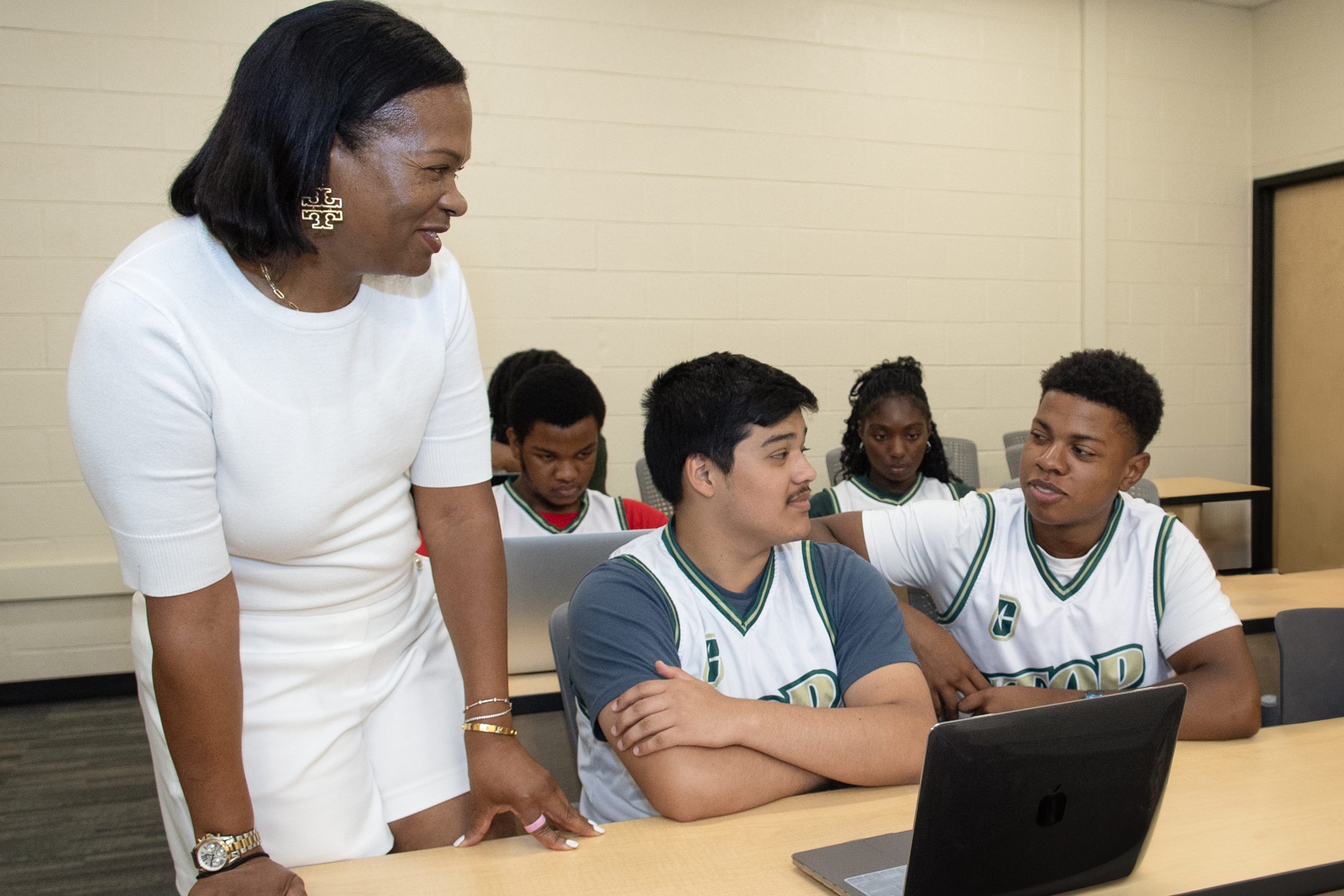 MAG_UTOP_Seberry_24031_8120_kpl (1) A woman interacts with students at a desk in a classroom, with laptops open.