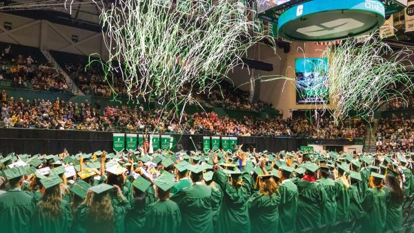 Graduates in green caps and gowns celebrate at a UNC Charlotte commencement ceremony inside a packed arena, as green and white streamers fall from the ceiling above a cheering crowd.