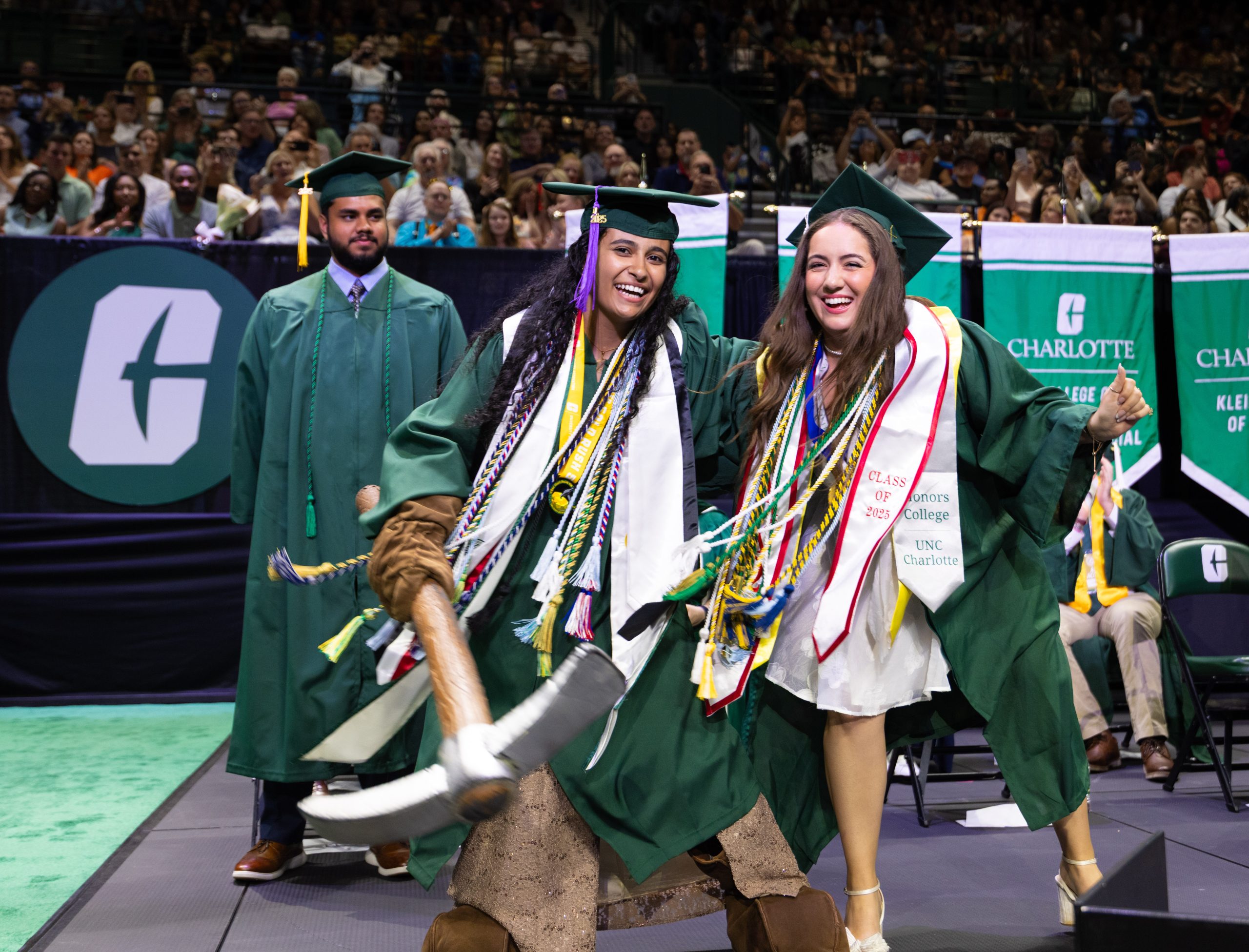 Two UNC Charlotte graduates in green gowns celebrate on stage, smiling and holding a ceremonial pickaxe.