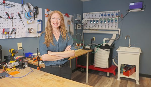 A woman stands confidently in a workshop with tools, electronics, and plumbing equipment organized behind her.