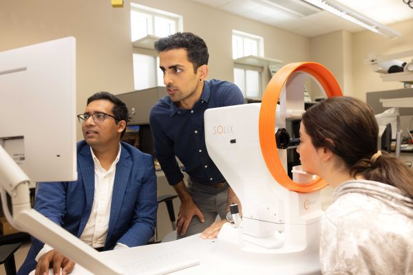Three people use a eye imaging device in a lab, with one person seated at a computer and another guiding a patient.
