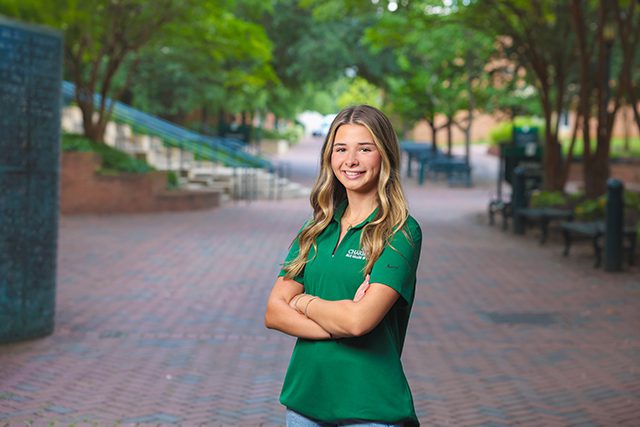 Student in green university polo stands confidently on a brick pathway lined with trees.