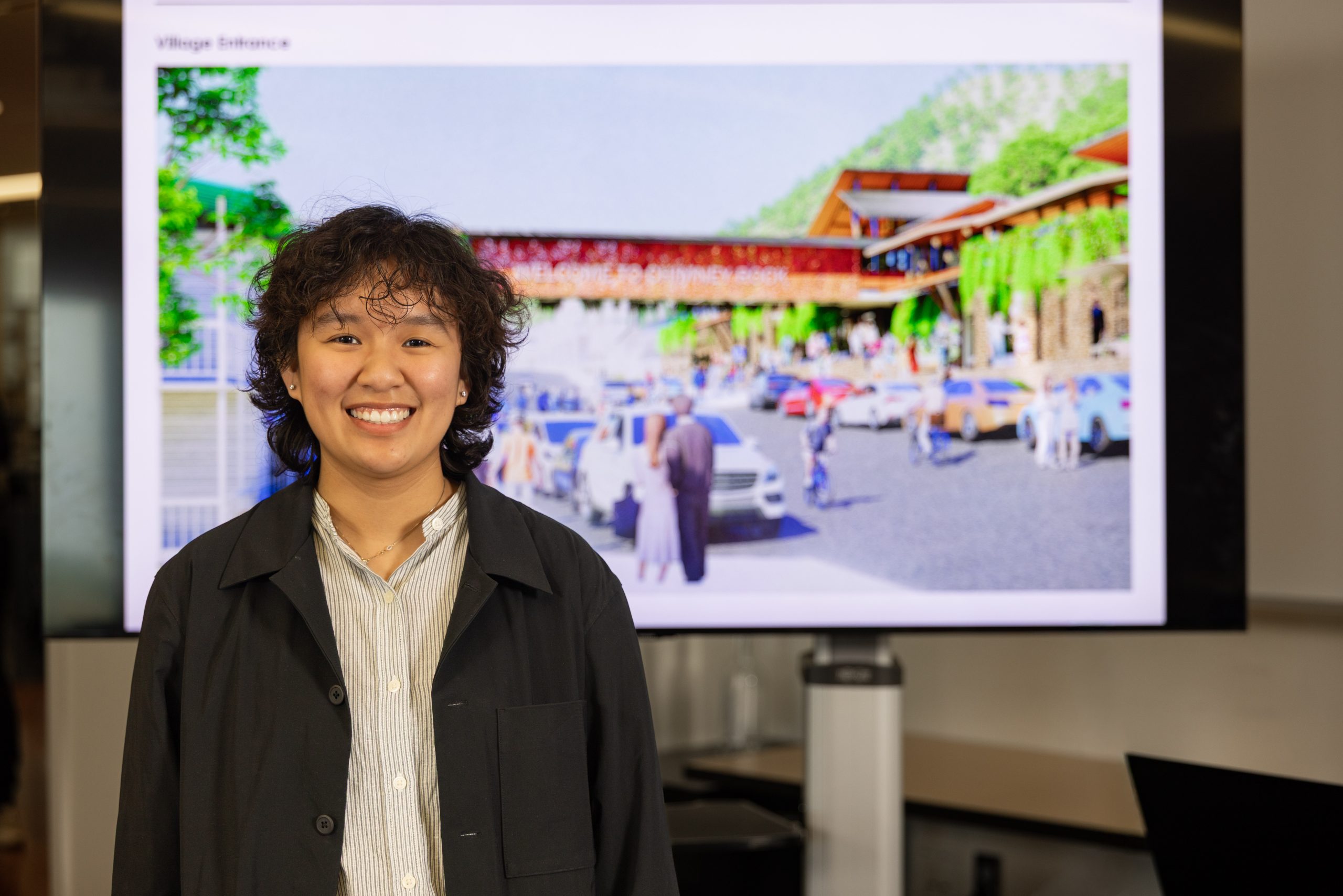 A student smiles while standing in front of a projected rendering of a village entrance and surrounding buildings.
