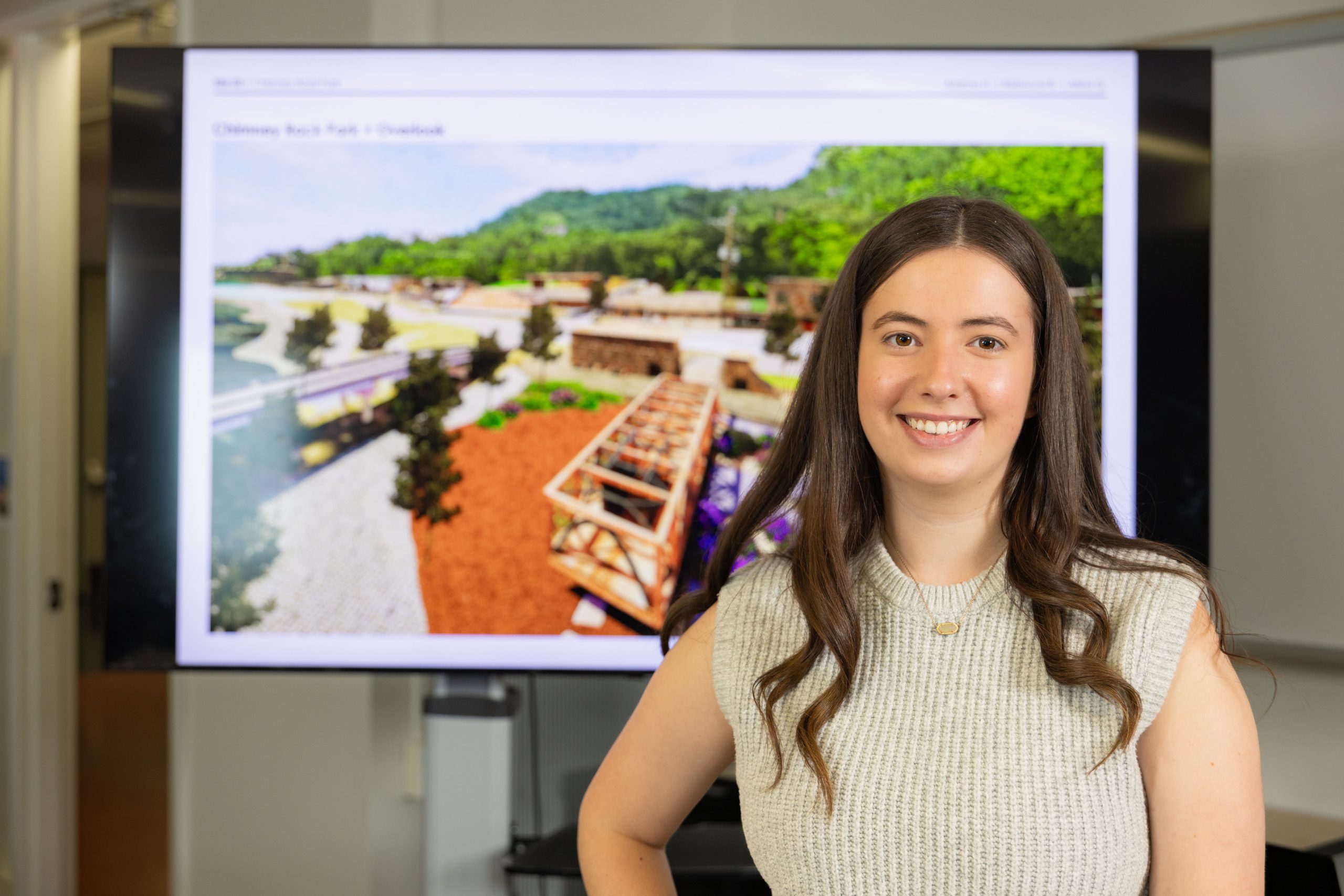 A student poses in front of a presentation screen displaying a rendering of a park overlook with walkways and landscaping.