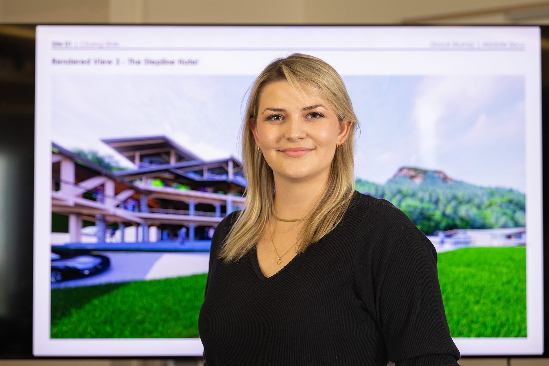 A student stands confidently in front of a presentation screen showing a rendering of a large building with green hills in the background.