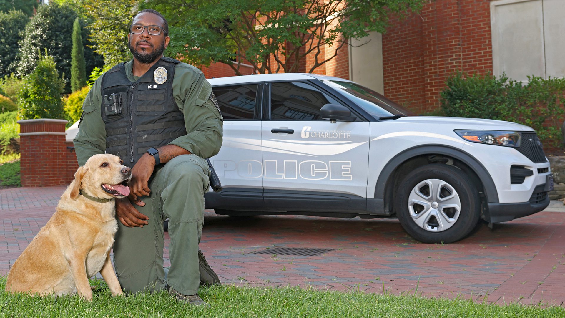 PPS_Group_Photos_25028_5783_kpl A police officer kneels beside a yellow Labrador Retriever in front of a campus police SUV.