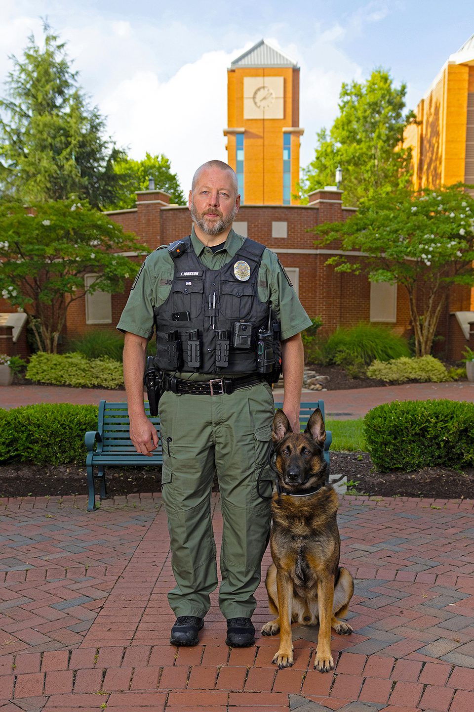 PPS_K9s_25001_5220_kpl A police officer stands with a German Shepherd sitting at his side in a brick courtyard with a clock tower in the background.