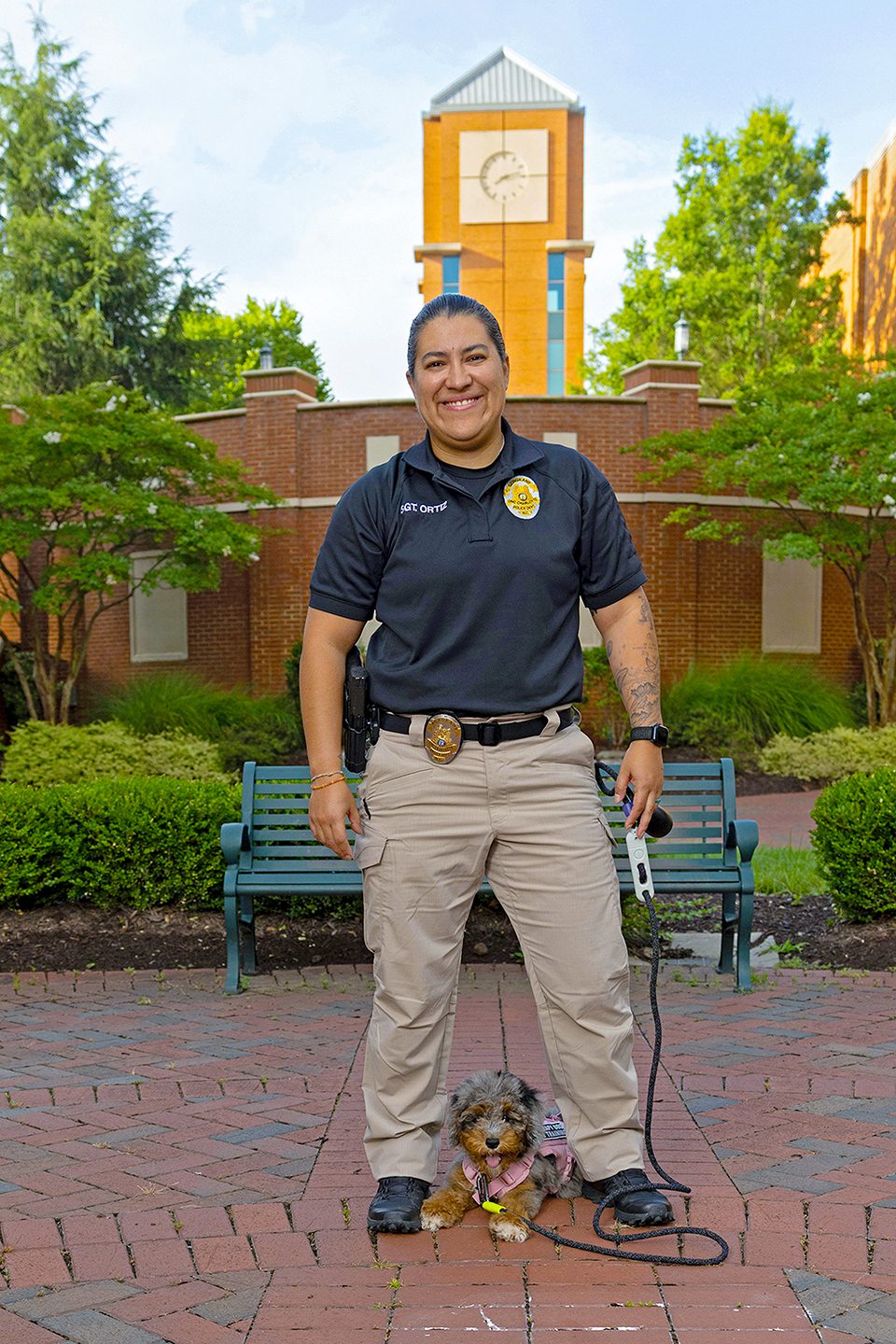 PPS_K9s_25002_5225_kpl A police officer smiles while standing with a small curly-haired dog on a leash in front of a campus clock tower.